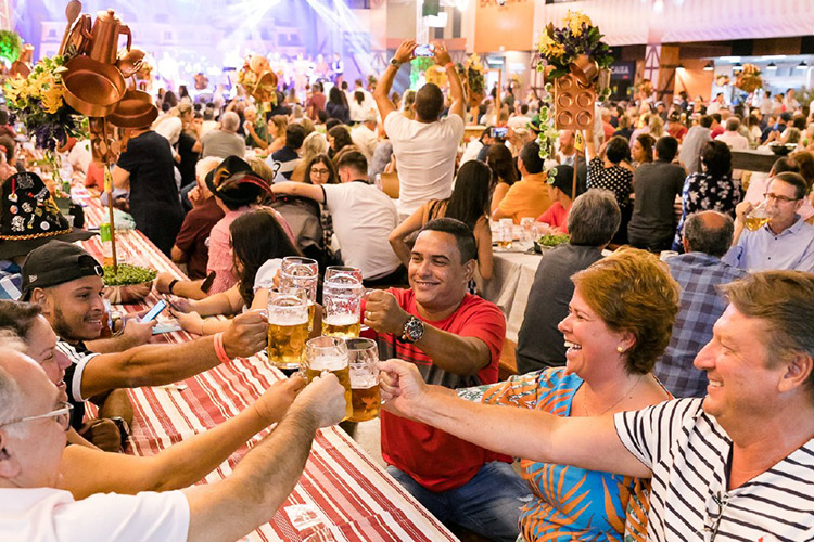 Na imagem vemos pessoas na Sommerfest sentadas à mesa, brindando com canecas de cerveja e sorrindo.
