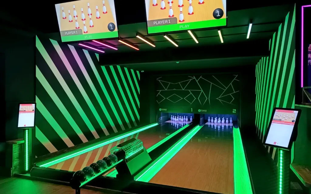 Ambiente de boliche com pista iluminada em verde e neon, telões exibindo partidas e pinos, além de controles laterais no placar, em arena de bowling