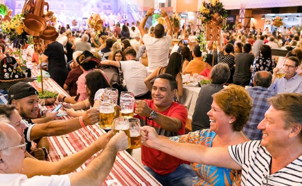 Na imagem vemos pessoas na Sommerfest sentadas à mesa, brindando com canecas de cerveja e sorrindo.
