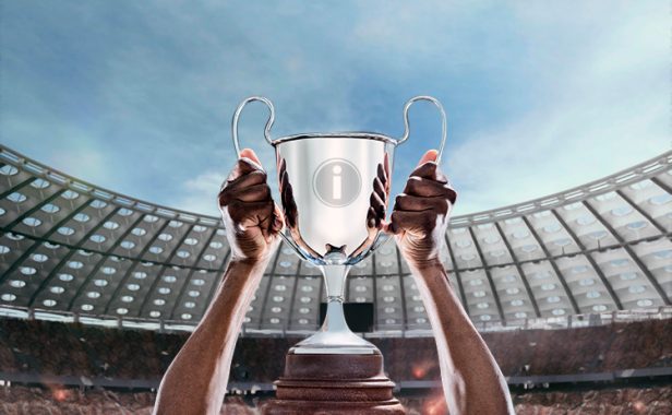 maiores-publicos-futebol-2022 Duas mãos levantando a uma taça com o logo da Imply no centro, em meio a um estádio todo iluminado e no topo o céu azul aparecendo.