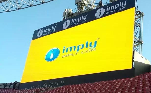 Mídia OOH em outdoor digital grande com a marca imprly, painel amarelo e logotipo i no topo, instalado em estrutura metálica com céu azul ao fundo.