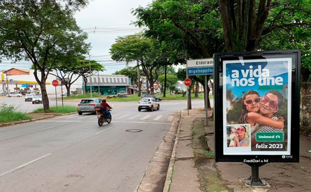 Ao lado direito da imagem podemos ver um painel fixo interativo com uma divulgação da Unimed. Localizado ao lado de uma via pública com árvores ao redor e veículos passando na faixa.