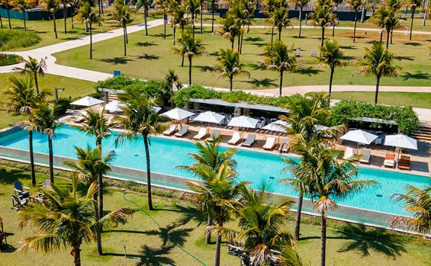 Visão externa do resort, mostrando a extensa piscina em meio a natureza com vários coqueiros ao redor e quiosques.