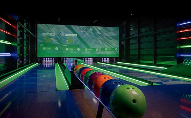 Na imagem é possível ver o ambiente do StrEAT Bowling de cima com as quatro pistas de boliche abertas, as bolas todas em posição no porta-bolas, luzes neons enfeitando as paredes laterais e canaletas. Ao fundo temos o painel ligado com interação e as pistas interativas com desenhos flutuantes.