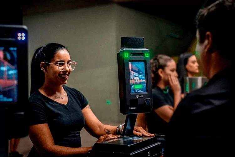 Mulher sorrindo diante de um dispositivo de identificação facial com leitor biométrico em ambiente de atendimento, enquanto outras pessoas observam ao fundo.