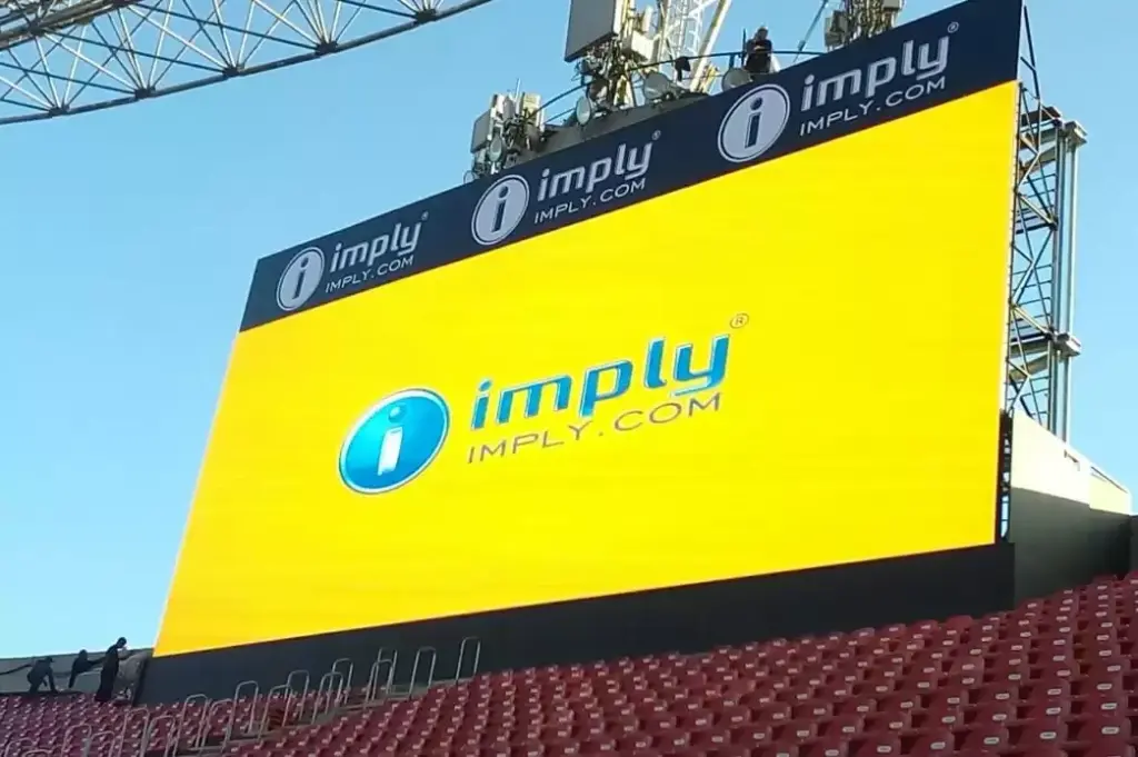 Mídia OOH em outdoor digital grande com a marca imprly, painel amarelo e logotipo i no topo, instalado em estrutura metálica com céu azul ao fundo.