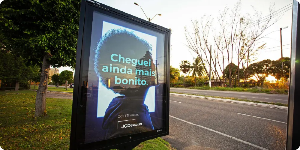 Mídia OOH em painel de rua com a mensagem “Cheguei ainda mais bonito” e marcações de publicidade, ao lado de uma avenida ao entardecer.