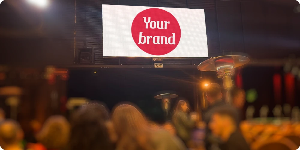 Mídia OOH em ambiente interno, com telão de LED iluminado exibindo o logo “Your Brand” em destaque ao fundo, cercado por pessoas desfocadas e luzes de palco