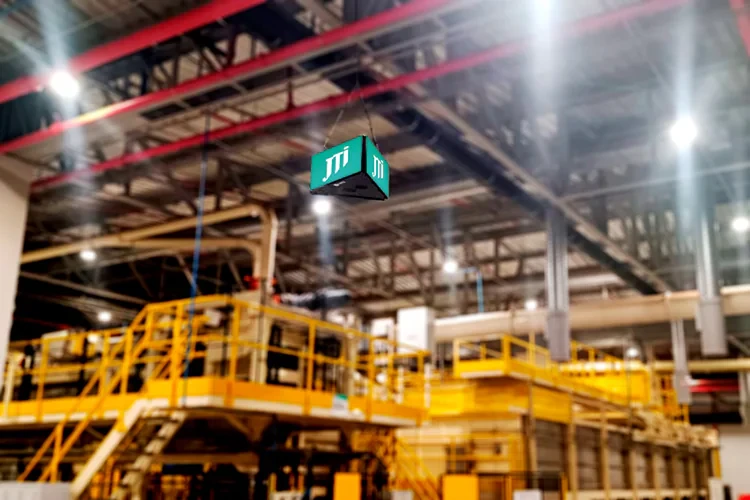 Planta industrial com maquinário e tubulações amarelas ao fundo, e um painel de LED indoor exibindo o logo JTI suspenso no alto do ambiente