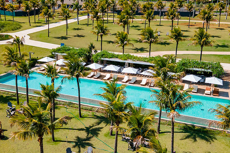 Visão externa do resort, mostrando a extensa piscina em meio a natureza com vários coqueiros ao redor e quiosques.