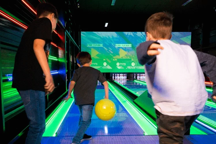 Pista de boliche iluminada com luzes verdes e neon, jogador com bola amarela prestes a arremessar, pessoas ao fundo e placar eletrônico ao longo das pistas