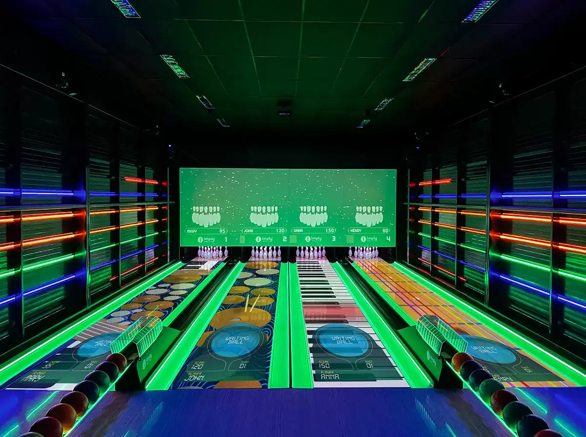 Vista interna de uma pista de boliche/bowling da Imply em ambiente futurista com iluminação neon verde, azul e vermelha, corredores iluminados e placar central com pinos, numerado para pontuação.