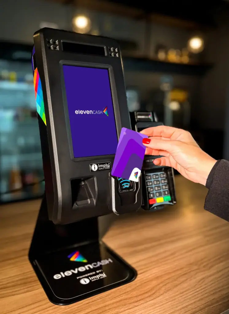 Terminal de pagamento cashless eleveNCash em checkout, com mão inserindo cartão roxo sem contato e visor exibindo “elevenCash”, em ambiente de loja